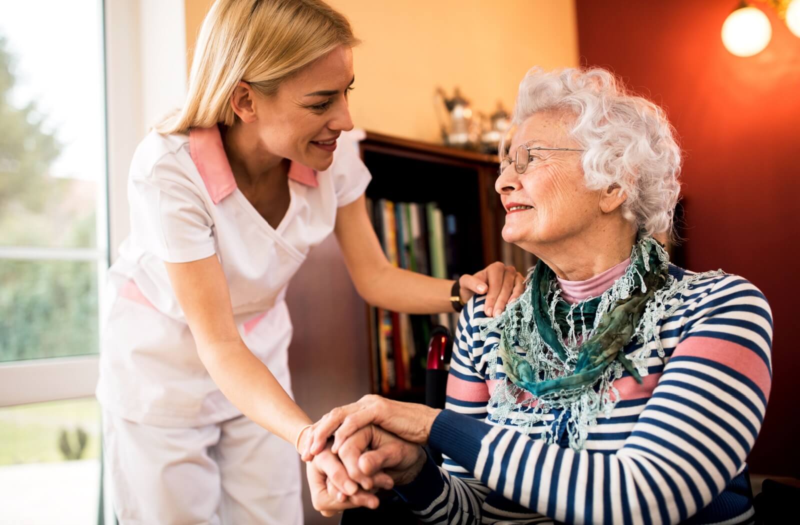 An older woman sitting down and holding hands with a nurse while they look and smile at each other