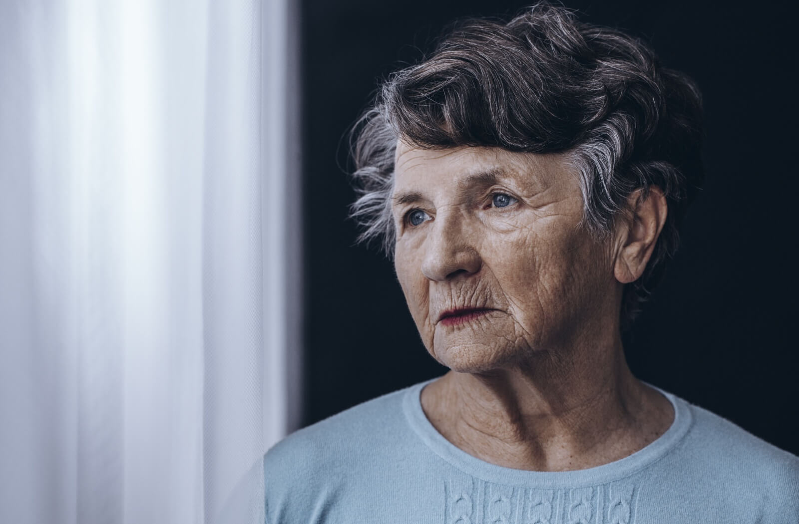 An older adult woman looking out the window with a serious expression