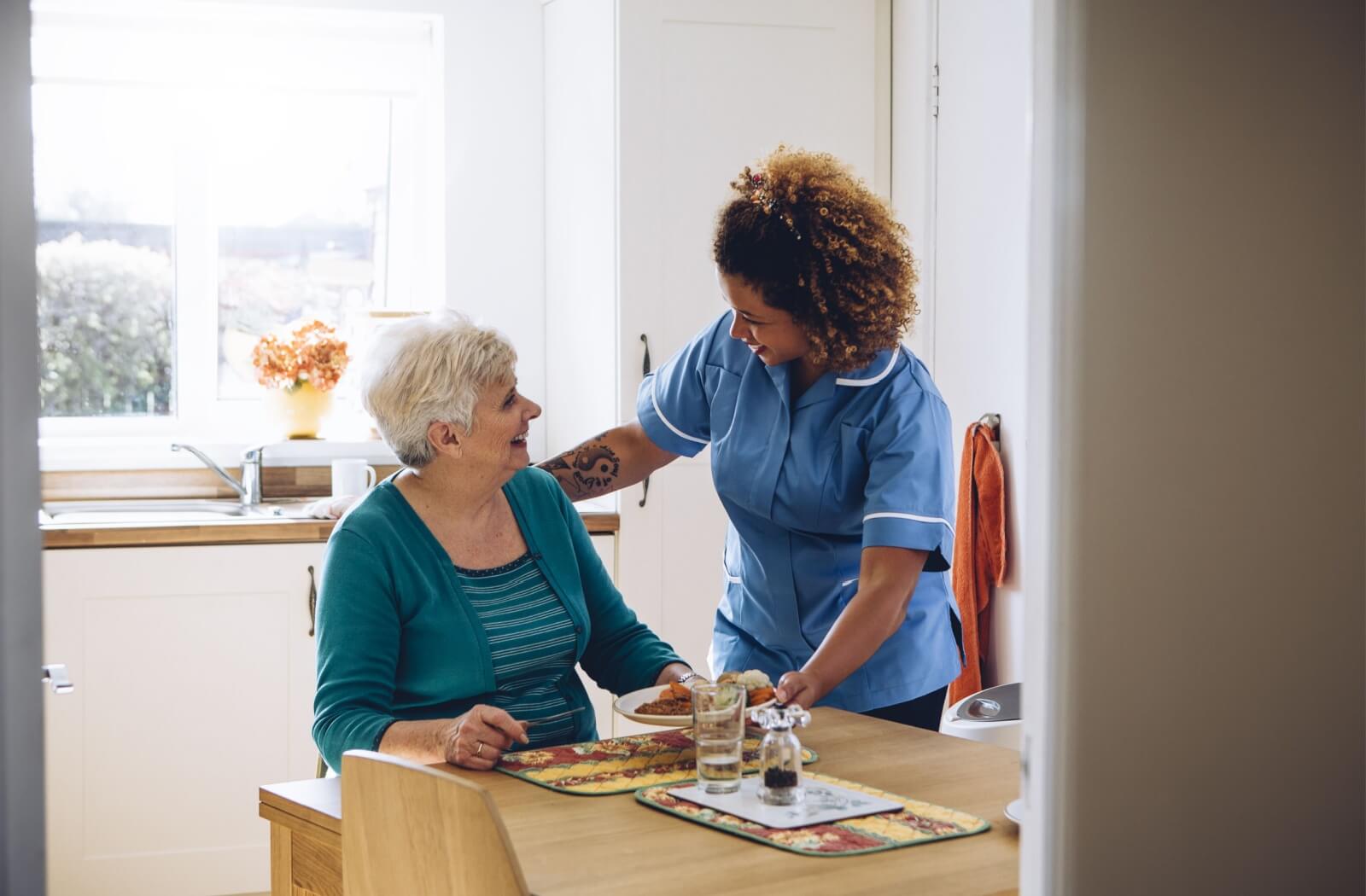 A female nurse resting her hand on an older woman's back while placing a plated meal in front of the senior and they smile and look at each other