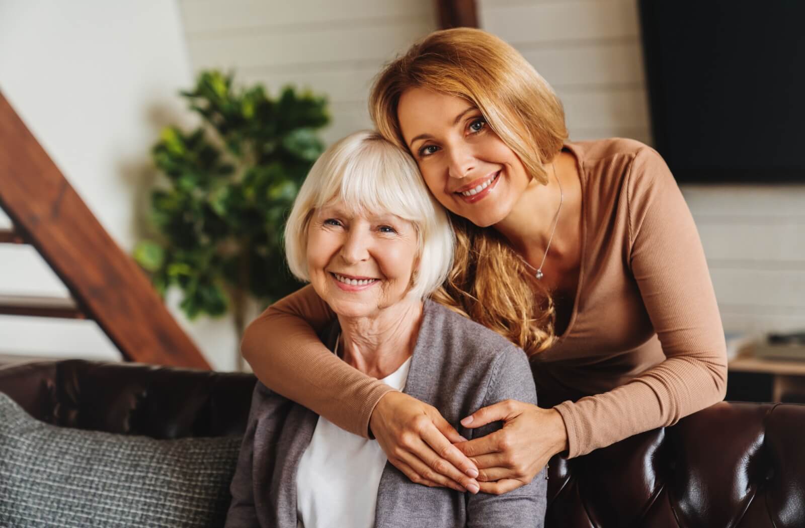 And older woman sitting on a couch while a younger woman hugs her from behind and they smile at the camera