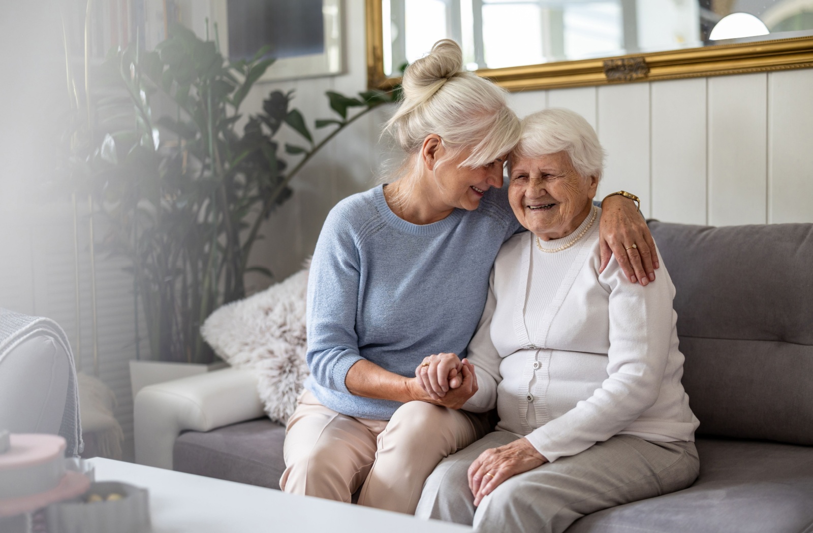 Smiling middle-aged daughter embracing her elderly mother on a couch, sharing a supportive moment.