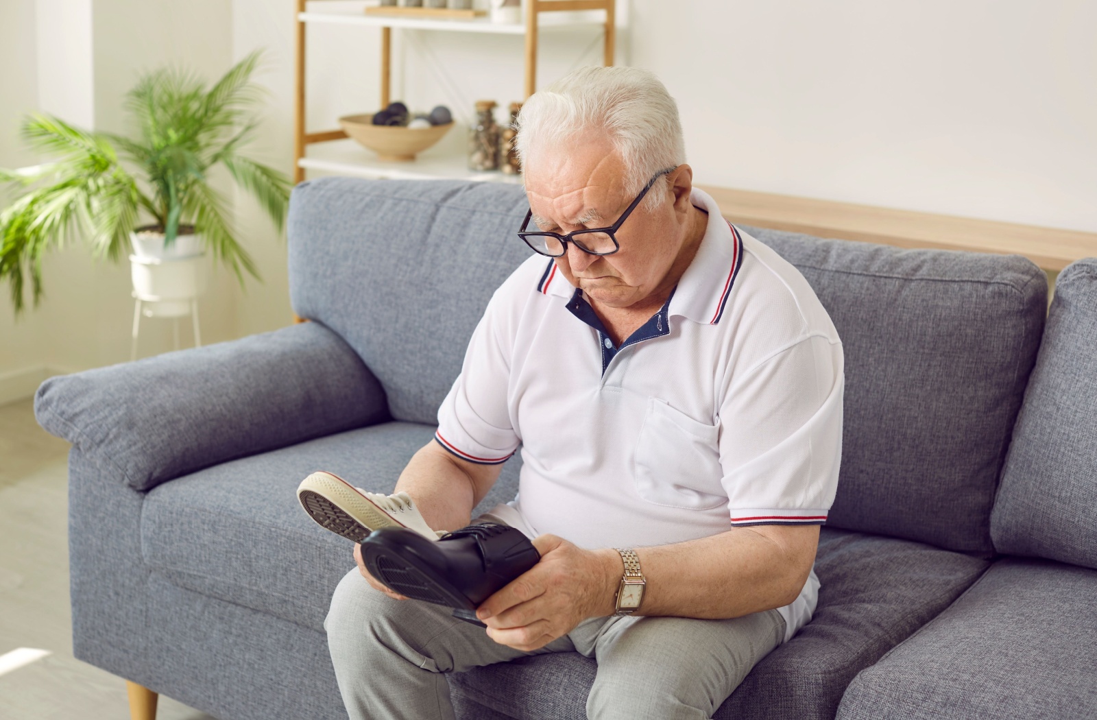 Elderly man sitting on a couch, looking confused as he examines a pair of shoes.