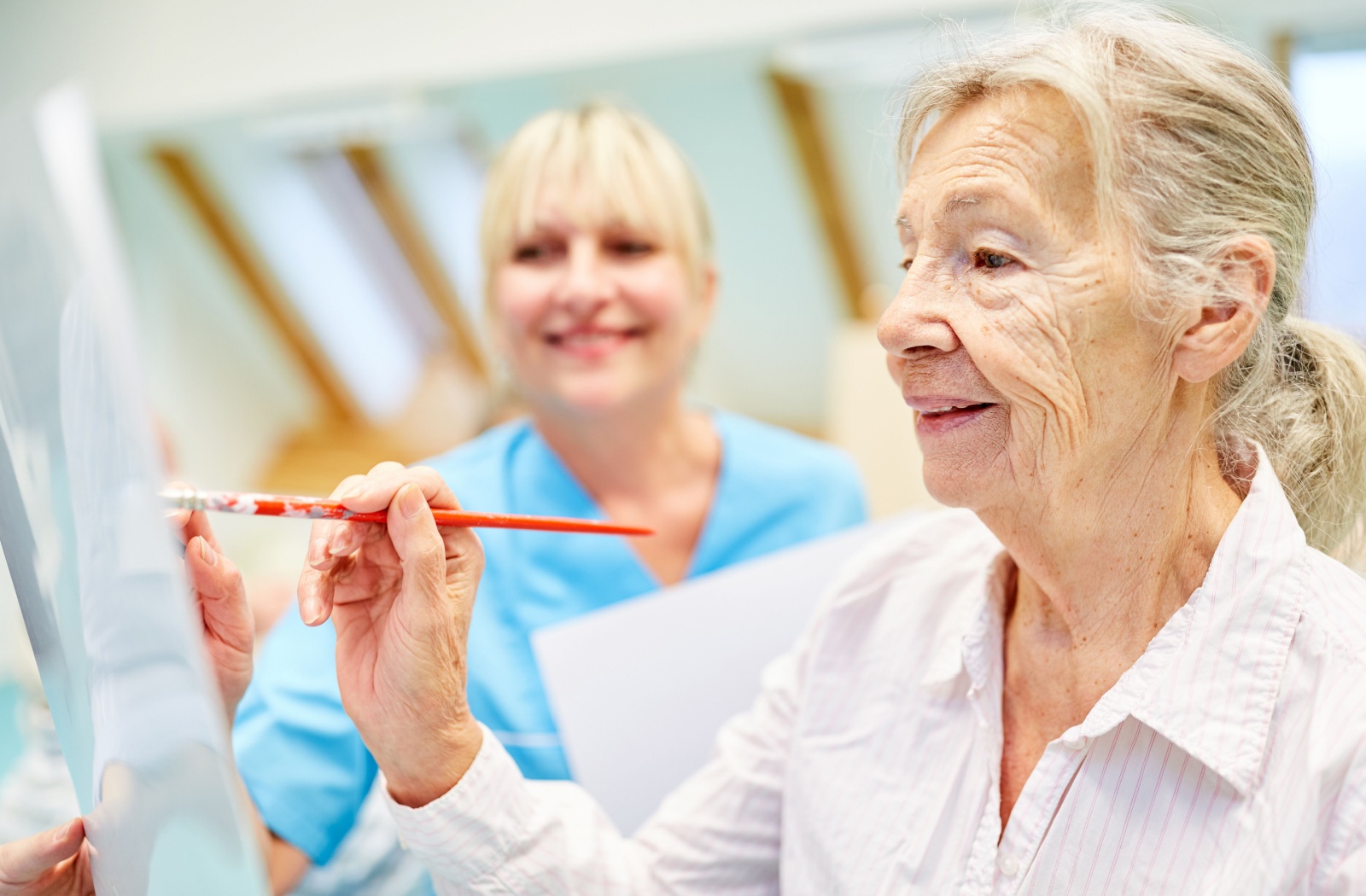 An older adult with dementia painting during an art class while a caregiver smiles in the background.