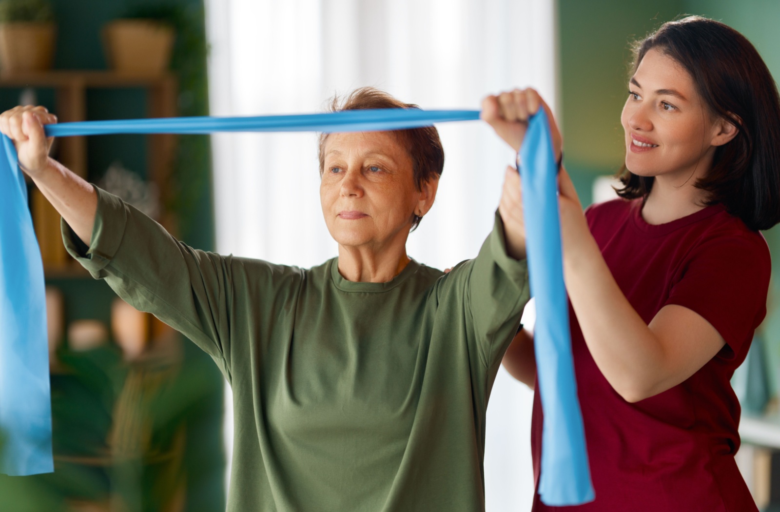 Senior in green shirt using resistance band with support from caregiver in maroon scrubs during an exercise