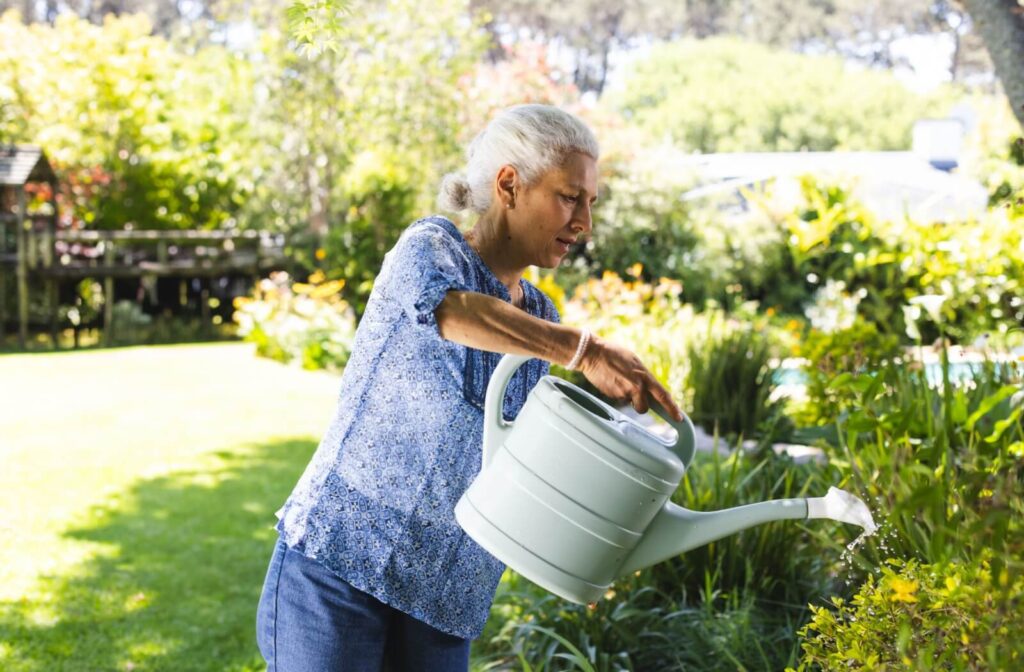 A senior wearing a blue floral top holds a large pale green watering can over a garden bed on a bright sunny day outdoors
