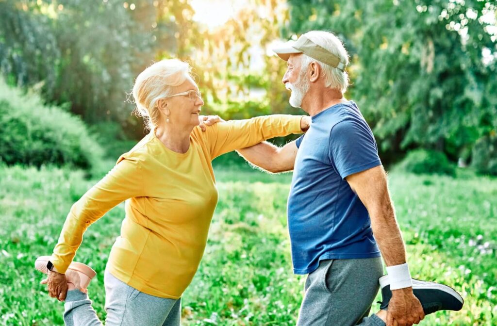 Two seniors stretching their legs together outside surrounded by lush greenery and offering each other support