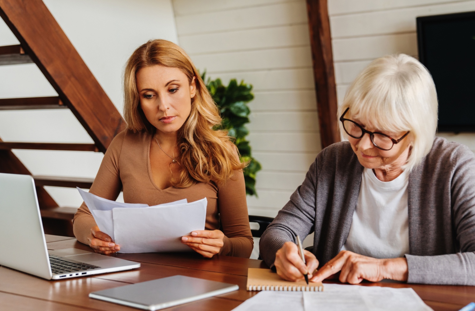 An adult child sits at a table with their older parent and helps them navigate legal documents