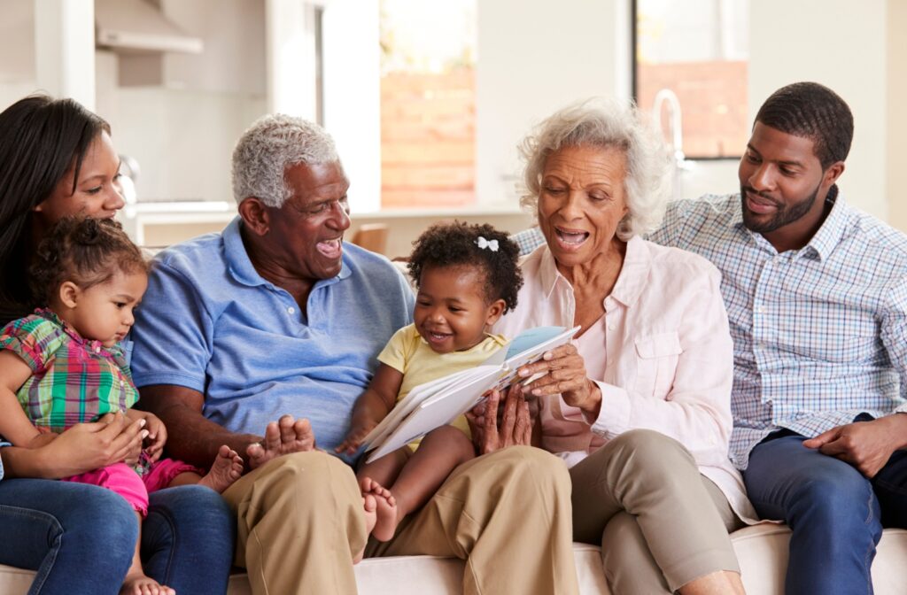 A senior couple sit on a couch and read with their grandchildren and adult children during a family visit to memory care