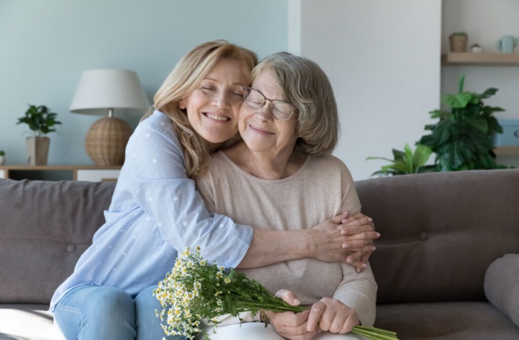 A middle-aged woman smiles while hugging her elderly mother tightly on a couch.