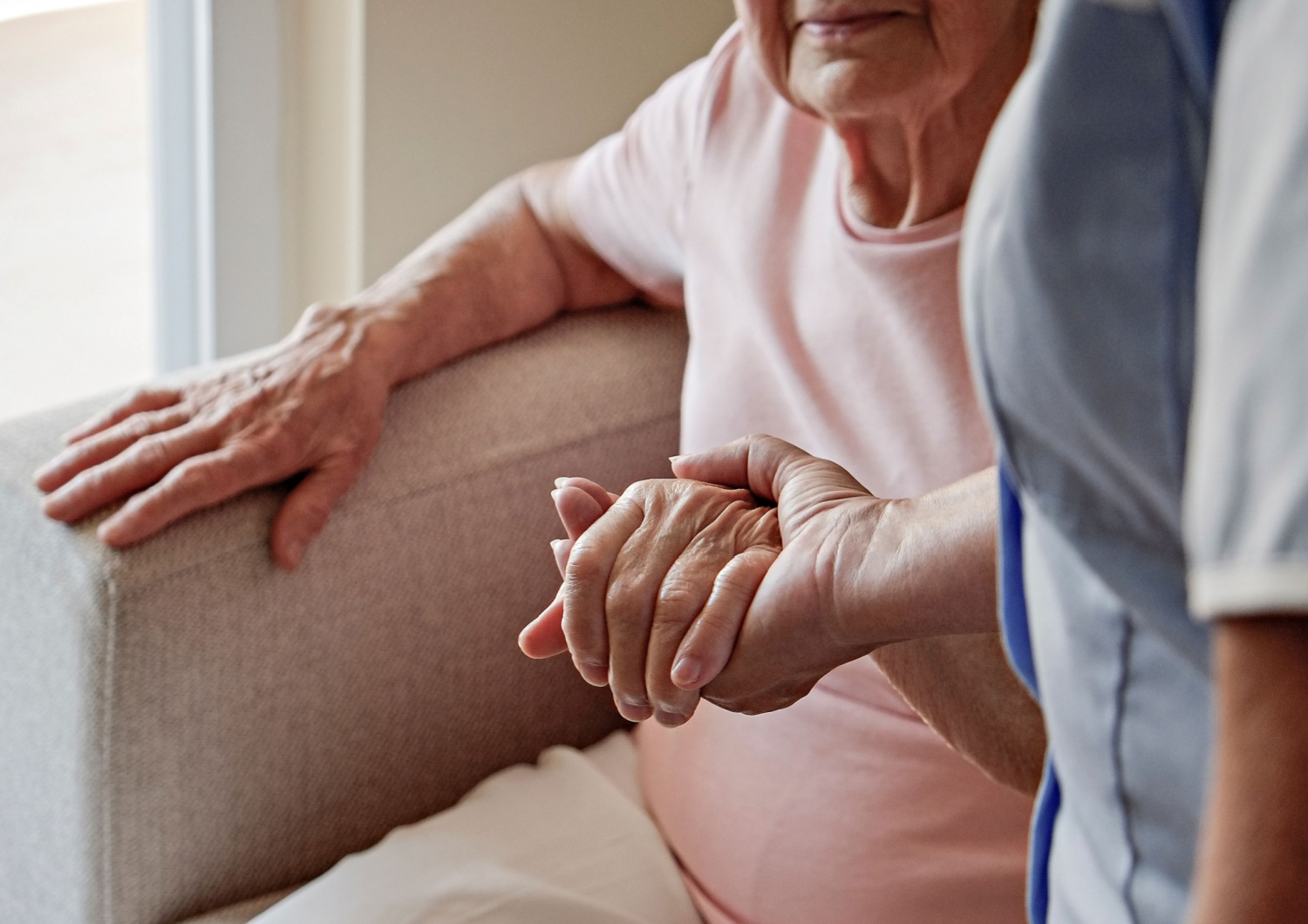 A caregiver holding an elderly woman's hand as she helps her stand up from a chair.